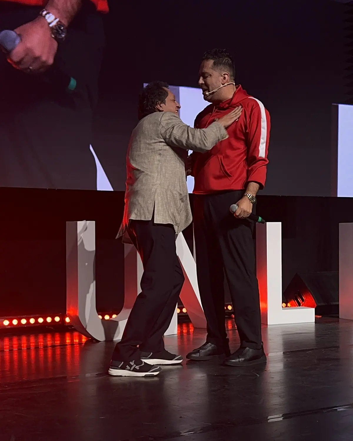Dos personas se abrazan en el escenario de Synergy Unlimited 2025, frente a las letras del evento